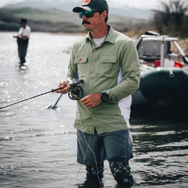 Man wearing the green camo Backcountry Skinz Airblitz Neoprene Wet Wading Pants while fishing in a body of water with a boat and another person in the background.