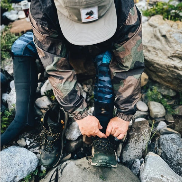 Person in camouflage pants and a cap tying wading boots over the grey neoprene Backcountry Skinz Z Drainz Guide Master Wading Sock on rocky terrain.