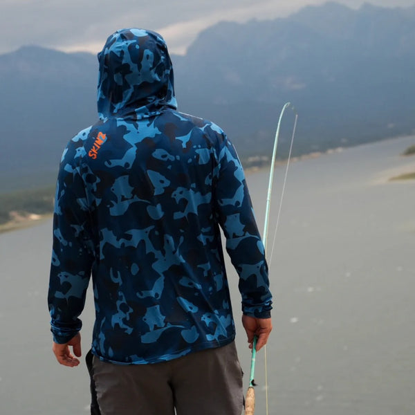 Person wearing blue camouflage Backcountry Skinz Men's Solarflexz UV Pro Sun Hoody, fly fishing rod in hand near a lake and mountains.