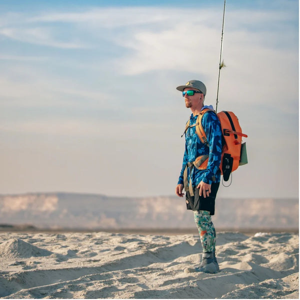 Person wearing blue camouflage Backcountry Skinz Men's Solarflexz UV Pro Sun Hoody, with a fly fishing rod and backpack, on a beach.