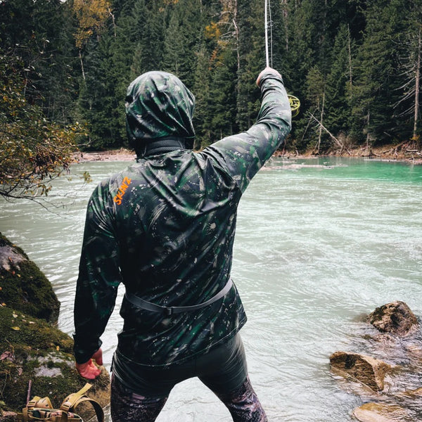 Person wearing green camouflage Backcountry Skinz Men's Solarflexz UV Pro Sun Hoody, fly fishing a stream in the forest.