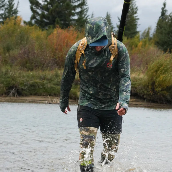 Person wearing green camouflage layers under the black Backcountry Skinz Men's Voltz Technical Shorts, walking through water with a backpack.