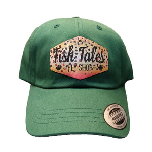 Green ball cap with brown trout 'Fish Tales Fly Shop' logo patch, on a white background.