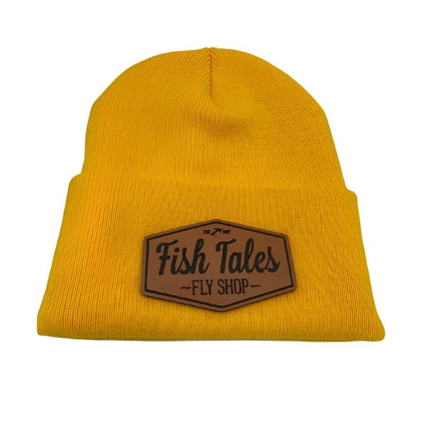 Front of a golden yellow knit beanie hat with a fish tales fly shop logo leather patch on a white background. 