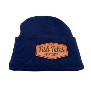 Front of a navy blue knit beanie hat with a fish tales fly shop logo leather patch on a white background. 