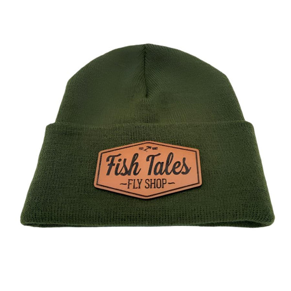 Front of an olive green knit beanie hat with a fish tales fly shop logo leather patch on a white background. 