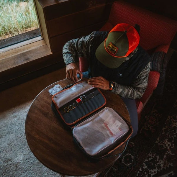 Person unpacking a green and grey Fishpond Backwater Fly Tying Kit containing fly tying materials and tools for travel, in a room by a window.
