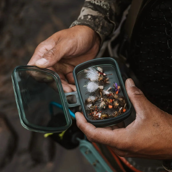 Person holding a smoke grey Fishpond Predator Fly Puck rectangular storage container full of flies on a rocky background.