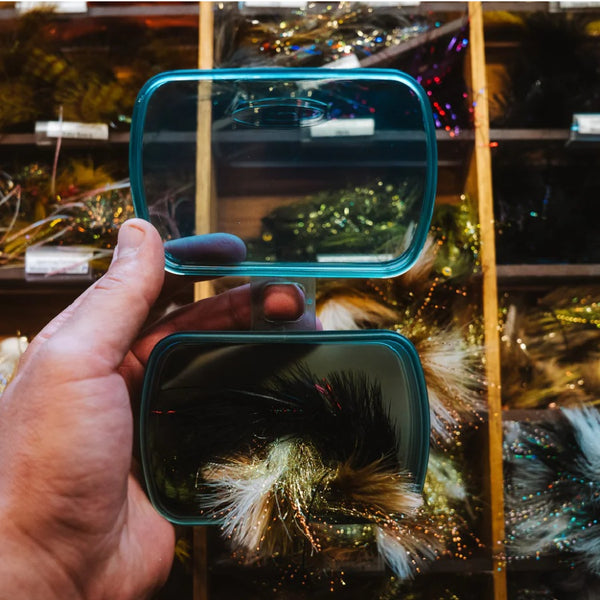 Person holding a smoke grey Fishpond Predator Fly Puck rectangular storage container full of flies over fly shop streamer bins.