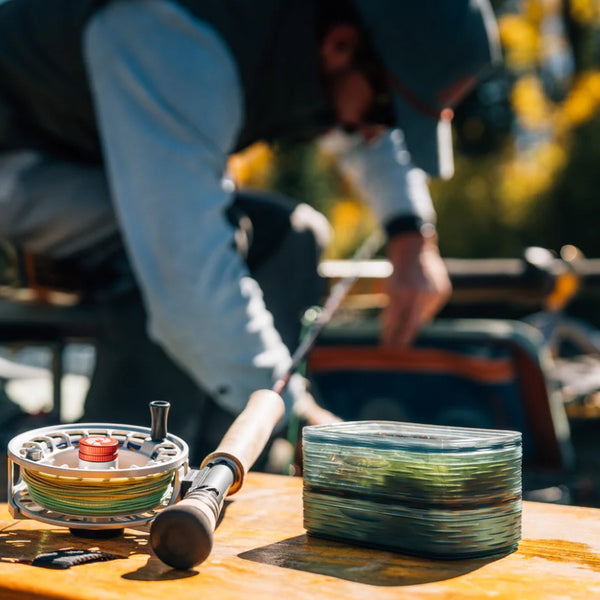 A stack of two smoke grey Fishpond Predator Fly Puck rectangular storage containers full of flies on a bench with a fly fishing rod and reel, with a person looking though a boat bag in the background.