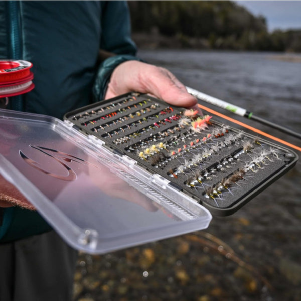 Person holding a Grey Fulling Mill stealth fly box with various flies by a body of water.