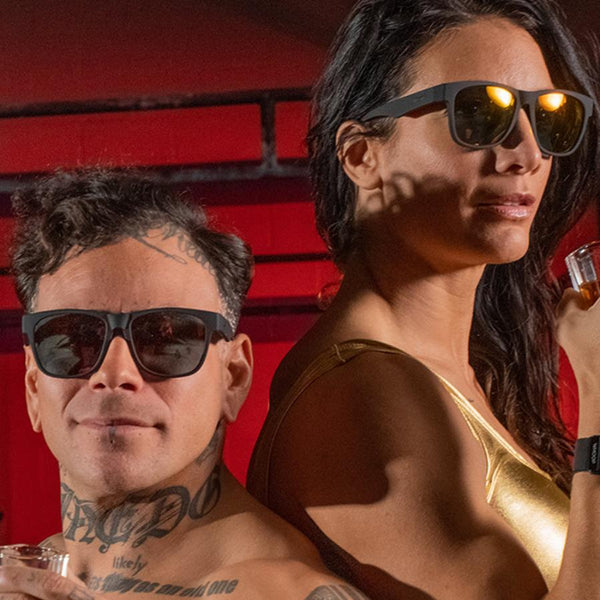 a man and a women wearing Goodr BFG Beelzebub's Bourbon Burpees Polarized Sunglasses on a red background