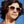 Person wearing beige Goodr BFG Big Pajama Llama Drama sunglasses with a gradient lens on a blue sky background.
