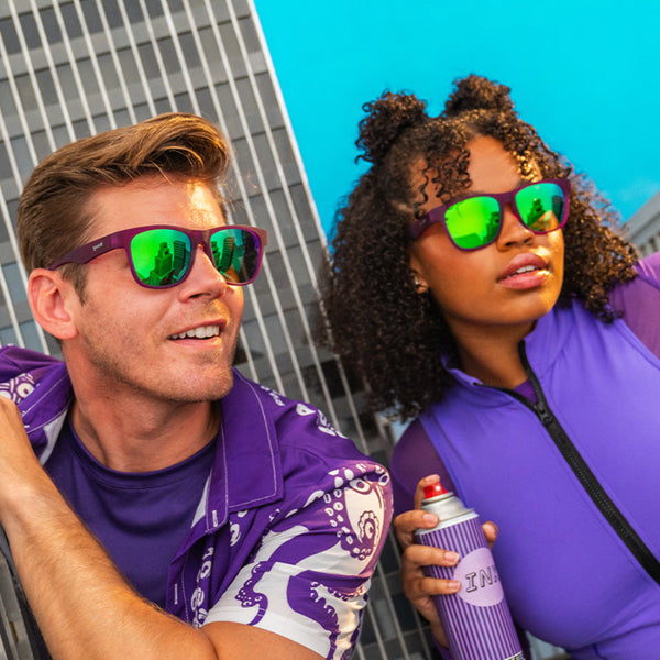 Two people wearing purple shirts and purple Goodr BFG Sunglasses with a cityscape background.