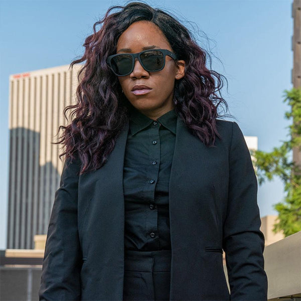 a woman in black clothing with long wavy dark hair wearing Goodr BFG Hooked On Onyx Polarized Sunglasses in front of a skyscraper.