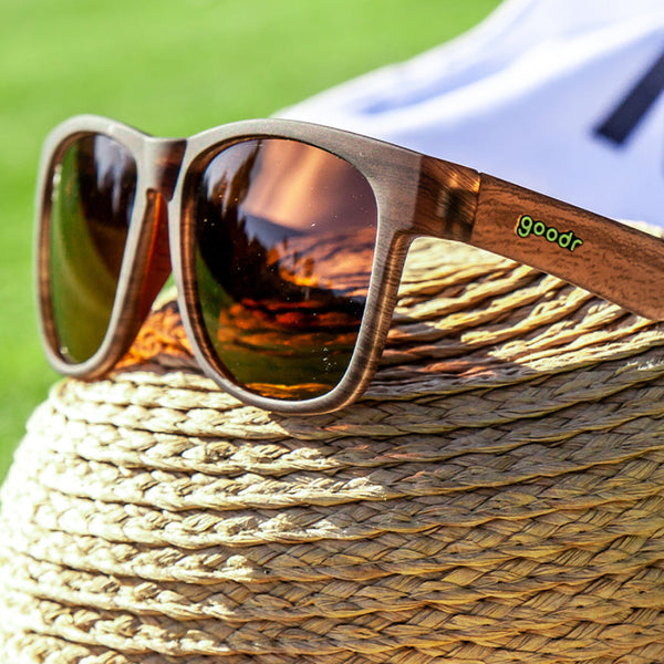 Goodr BFG wood grain frame sunglasses with reflective lenses on a straw hat against a blurred natural background.