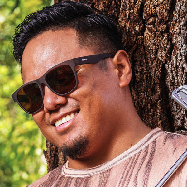 Man wearing Goodr BFG sunglasses leaning against a tree with a blurred green background.