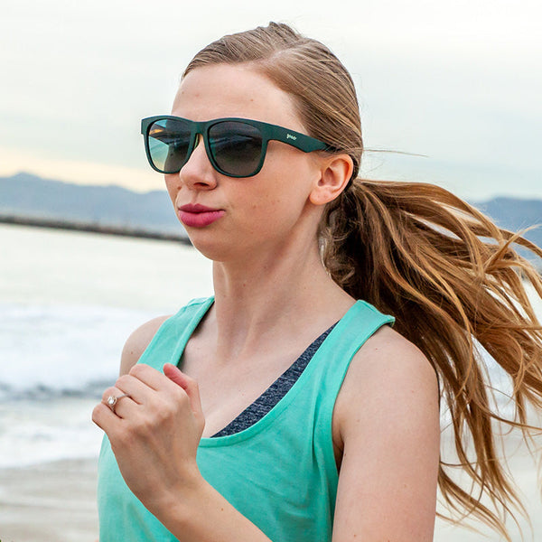 a woman with a long blonde ponytail running on the beach wearing goodr bfg mint julep electroshocks polarized sunglasses and a teal blue tank top