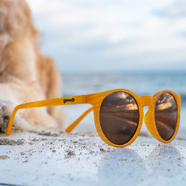 Wood patterned Goodr Circle G Bodhi's Ultimate Ride sunglasses with brown lenses in a beach setting with an ocean background.