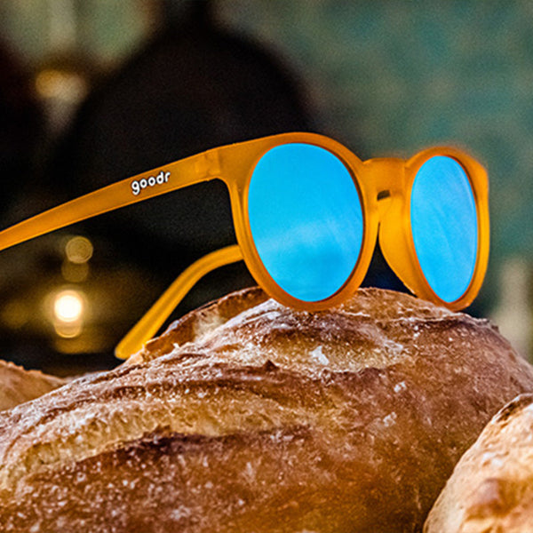 Orange Goodr Circle G sunglasses with blue lenses on a loaf of bread against a blurred background.