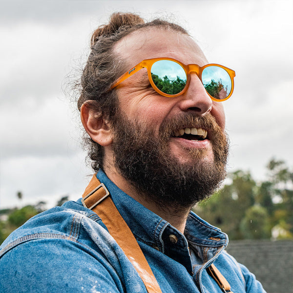 Person with bun and beard wearing Orange Goodr Circle G sunglasses with blue lenses on a blurred natural background.