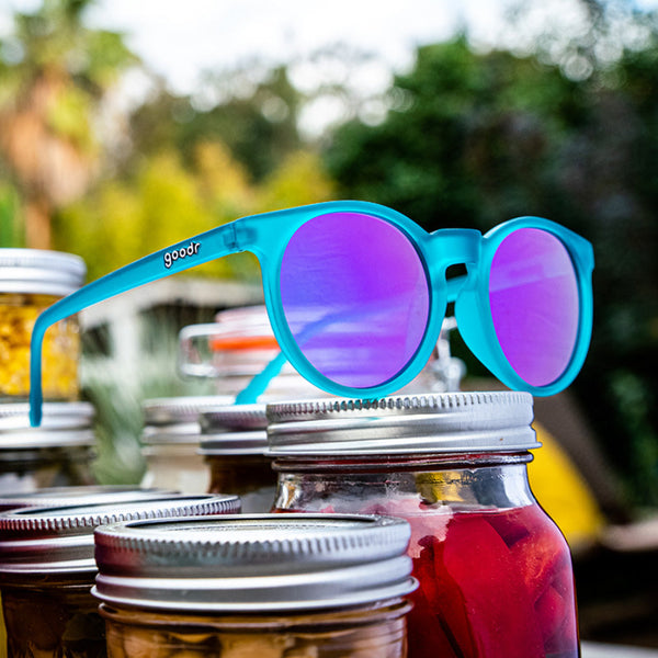 Teal framed Goodr Circle G I Pickled These Myself Sunglasses with lilac lenses sitting on pickle jars with a blurred nature background.