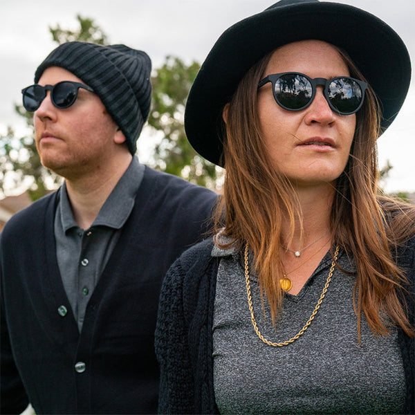 Two people wearing black Goodr Circle G sunglasses and hats outdoors with trees in the background.