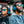 Two people wearing Goodr Circle G Sunglasses with blue lenses and black frame in front of a wall of vinyl records.