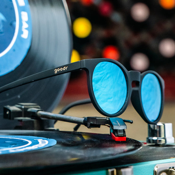 Goodr Circle G Sunglasses with blue lenses and black frame sitting on a record player on a blurred background.