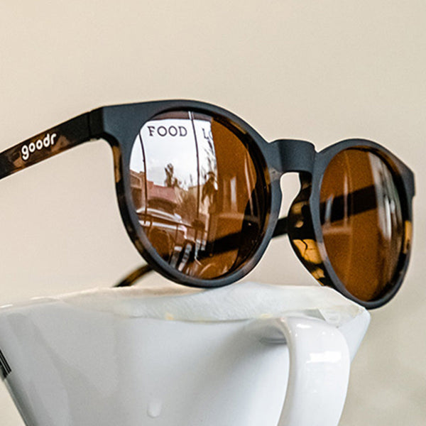 Coffee cup with Goodr Circle G Nine Dollar Pour Over Sunglasses with tortoiseshell frame over an off-white background.