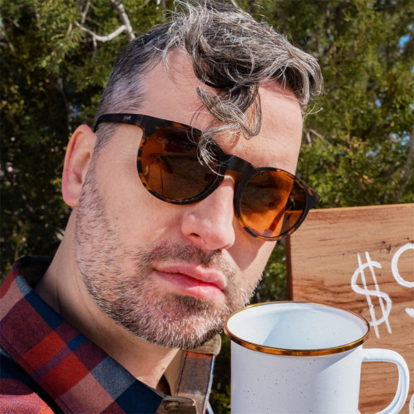 Person holding a coffee cup wearing a plaid shirt and Goodr Circle G Nine Dollar Pour Over Sunglasses with tortoiseshell frame with trees in the background.