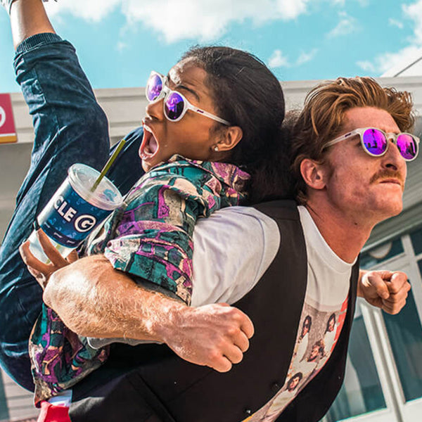 Two people wearing Goodr Strange Things are Afoot at the Circle G Sunglasses with white frame and pink lenses with clear sky and buildings in the background.