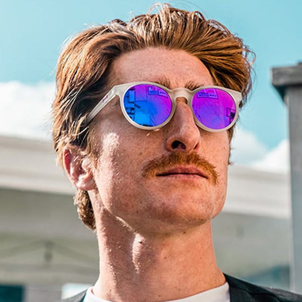 Person wearing Goodr Strange Things are Afoot at the Circle G Sunglasses with white frame and pink lenses with clear sky and buildings in the background.