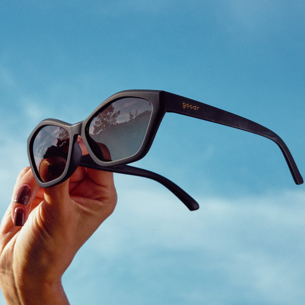 Hand holding black Goodr Deco G sunglasses against a blue sky.