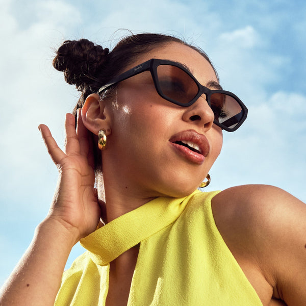 Woman wearing black framed Goodr Deco G Sing in Reverb Sunglasses and a yellow top against a blue sky