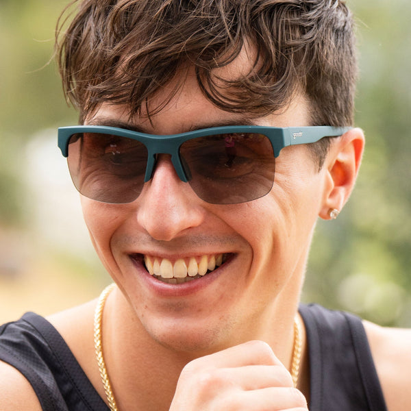 Man wearing Goodr Flex G Lawn Mower Drag Race Sunglasses with green frame and brown gradient lens on a blurred natural background.