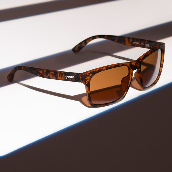Goodr Grand G Sunglasses with tortoiseshell frame and brown lenses on a shadowed surface.