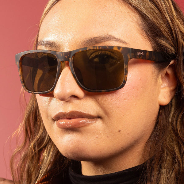 Close-up of a person wearing Goodr Grand G tortoiseshell sunglasses with a pink background