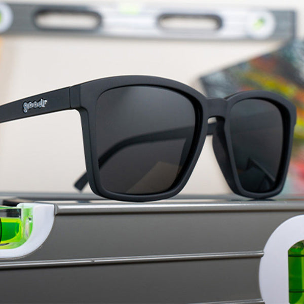 A pair of matte black Goodr LFG sunglasses on a level with a blurred background.