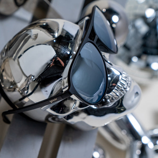 Black Goodr MACH G Add The Chrome Package aviator sunglasses sitting on a silver skull on a blurred grey background.