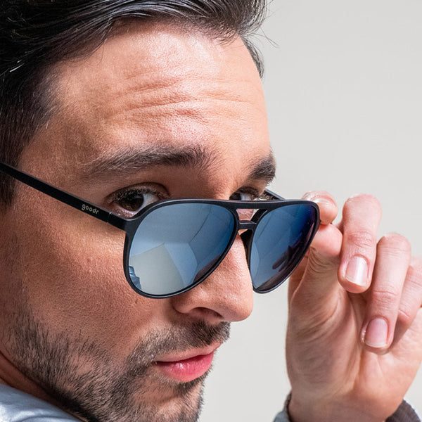 Person wearing black Goodr MACH G Add The Chrome Package aviator sunglasses with silver reflective lenses on a neutral background.