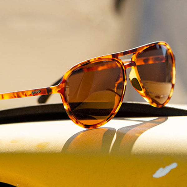 Goodr Mach G Aviator Tortoiseshell sunglasses on a reflective surface with a blurred background.