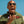 Man in a green flight suit with patches, wearing Green Goodr Mach G Buzzed on the Tower sunglasses, against a clear blue sky.