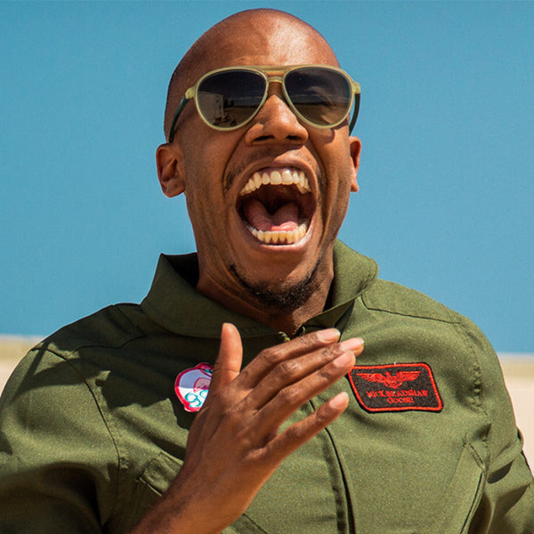 Man in a green flight suit with patches, wearing Green Goodr Mach G Buzzed on the Tower sunglasses, against a clear blue sky.
