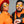 Two people wearing black and orange outfits and Goodr MACH G Call Me Tarmac Daddy sunglasses against a gray background.
