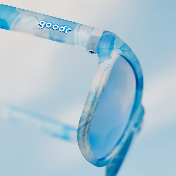 Close up of blue and white Goodr OG Binge Watching the Sky sunglasses with blue lenses on a light blue background.