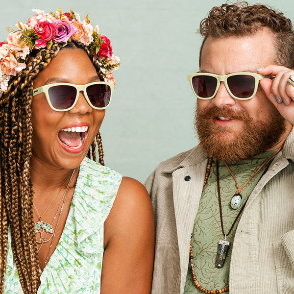 Two people wearing Green Goodr OG Dawn Of A New Sage sunglasses with purple gradient lenses on a white background on a light green background.