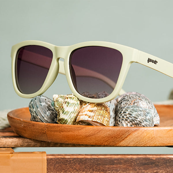 Green Goodr OG Dawn Of A New Sage sunglasses with purple gradient lenses on wooden shelf with seashells.