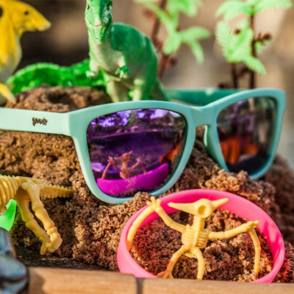 Toy skeleton and dinosaur figures in sand with the Goodr OG Electric Dinotopia Carnival sunglasses with a beachy background.
