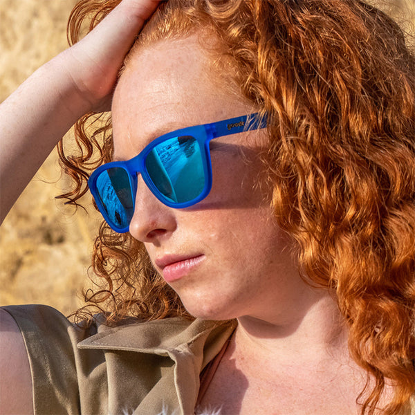 Person with red hair wearing Blue Goodr OG Falkor's Fever Dream Sunglasses on a blurred natural background.